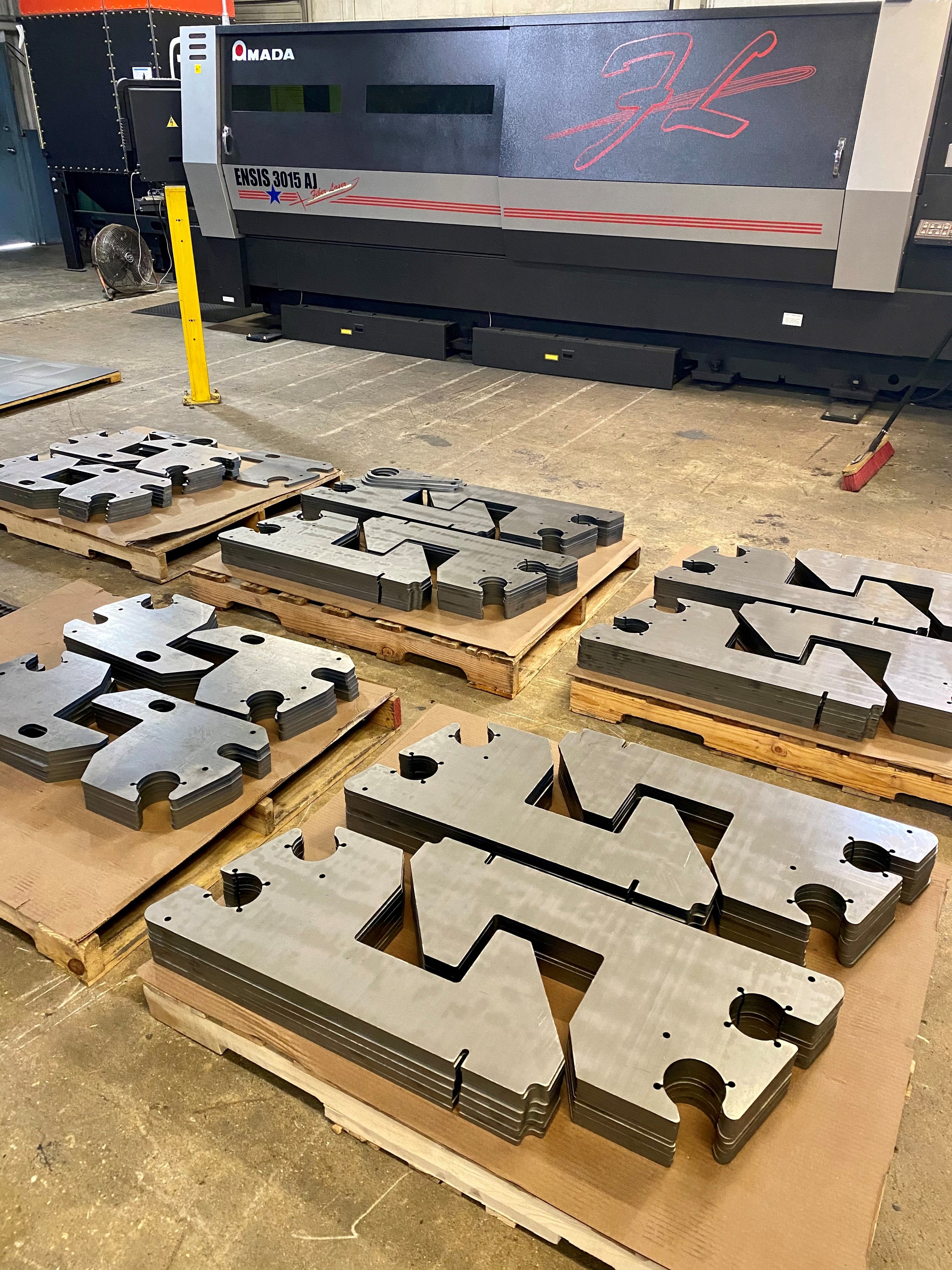 OP Machine and Fabrication laser cut sheet metal components staged on wooden pallets with an Ensis3015 3kw laser machine in the background.